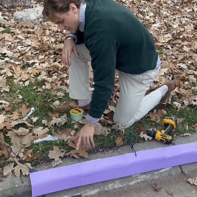 Josh kneeling at a residential curb, fitting the first Bizzi prototype strip along the gutter line, with a drill and tape beside him.